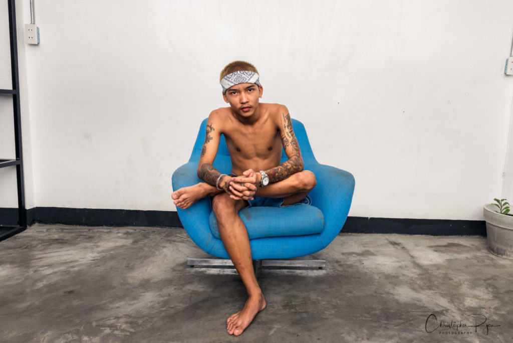 sexy teen boy with no shirt wearing a white headband sitting in blue chair barefoot with tattoos