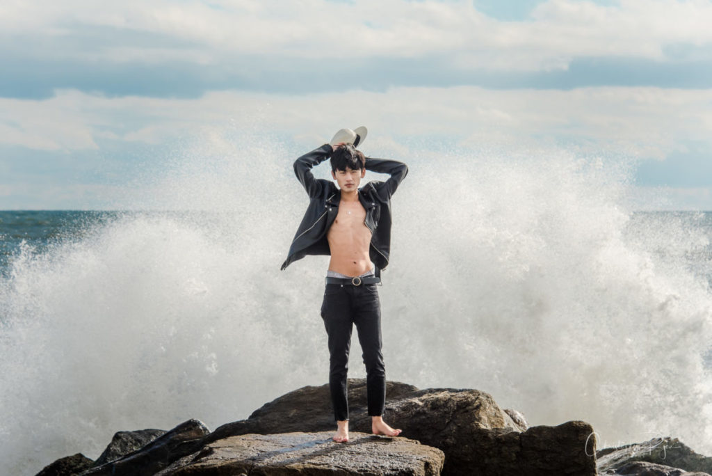 asian boy standing on rock jetty with huge wave behind him