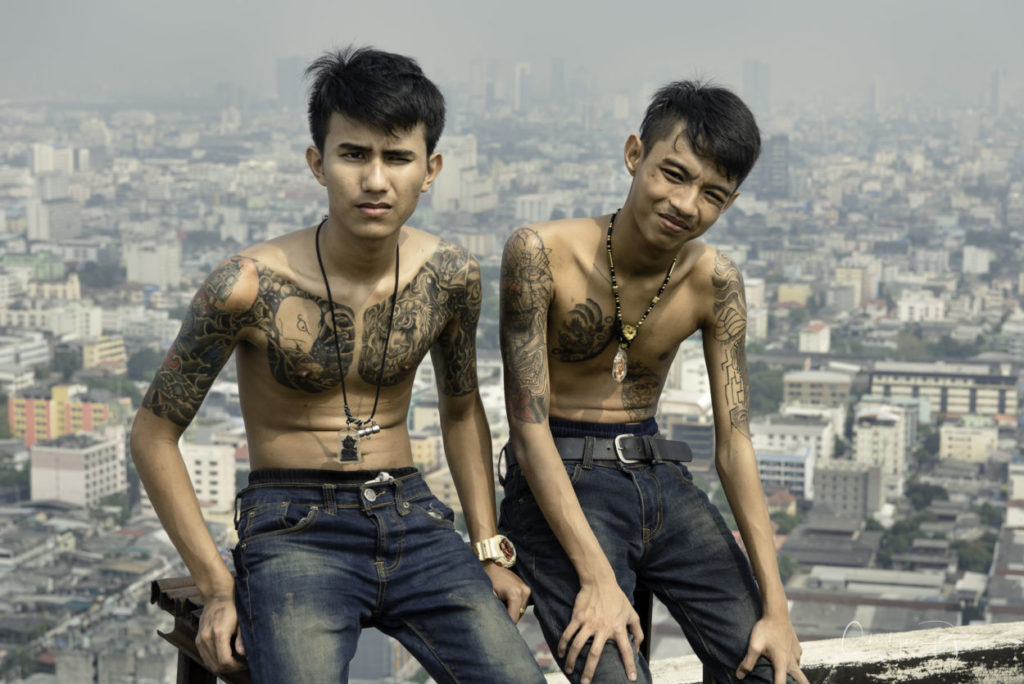 two shirtless boys in bluejeans and tattoos on top of a skyscraper